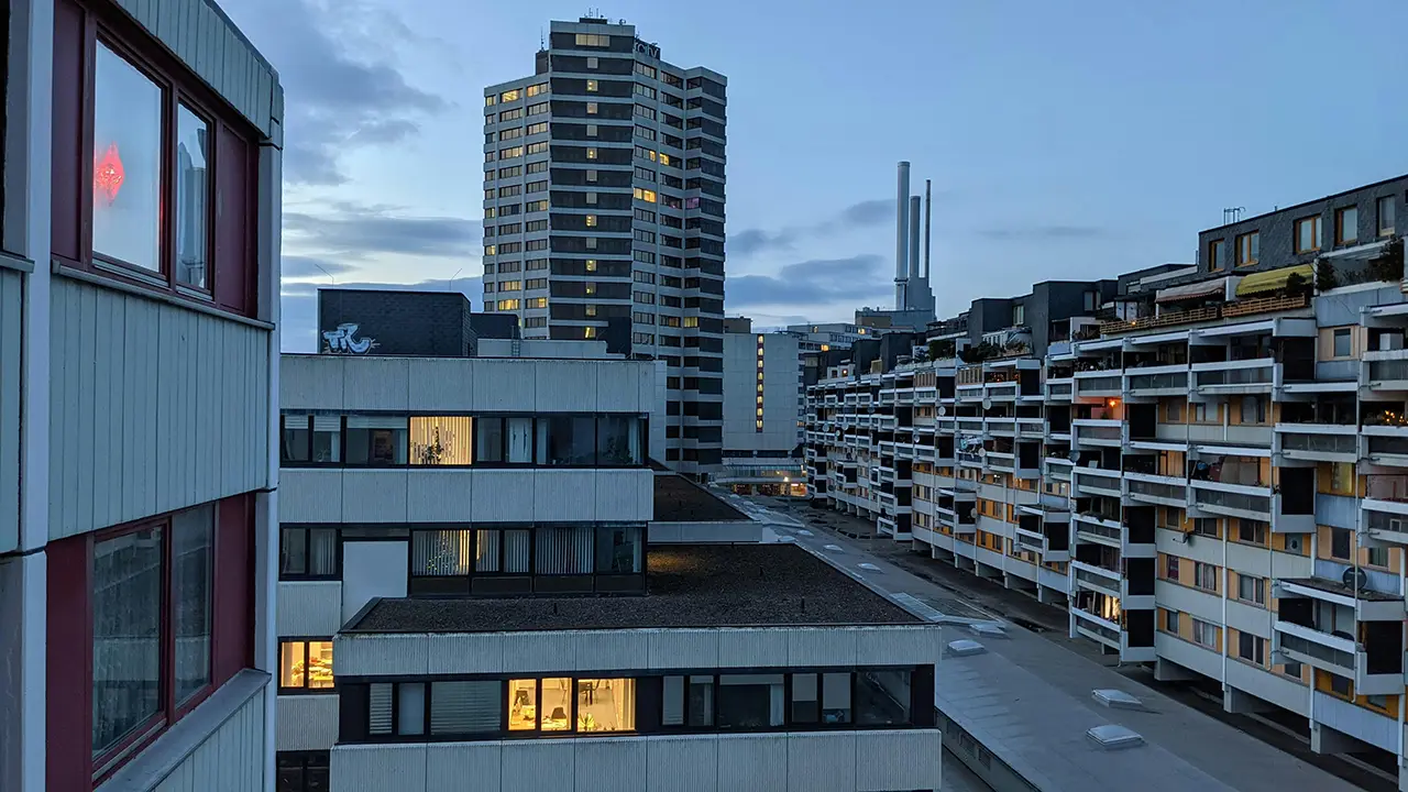 Ansicht des Ihme-Zentrums in Hannover mit dem Heizkraftwerk ‚Drei warme Brüder‘ im Hintergrund, aufgenommen in der Abenddämmerung mit erleuchteten Fenstern.