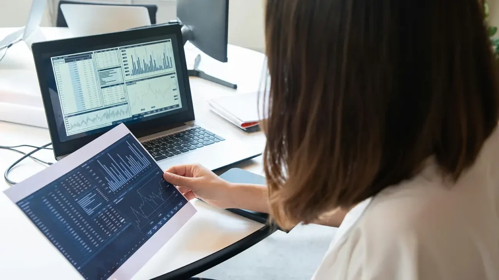 Frau betrachtet Diagramme und Statistiken auf Laptop und Ausdruck – Symbol für Analyse von Webdesign und Suchmaschinenoptimierung