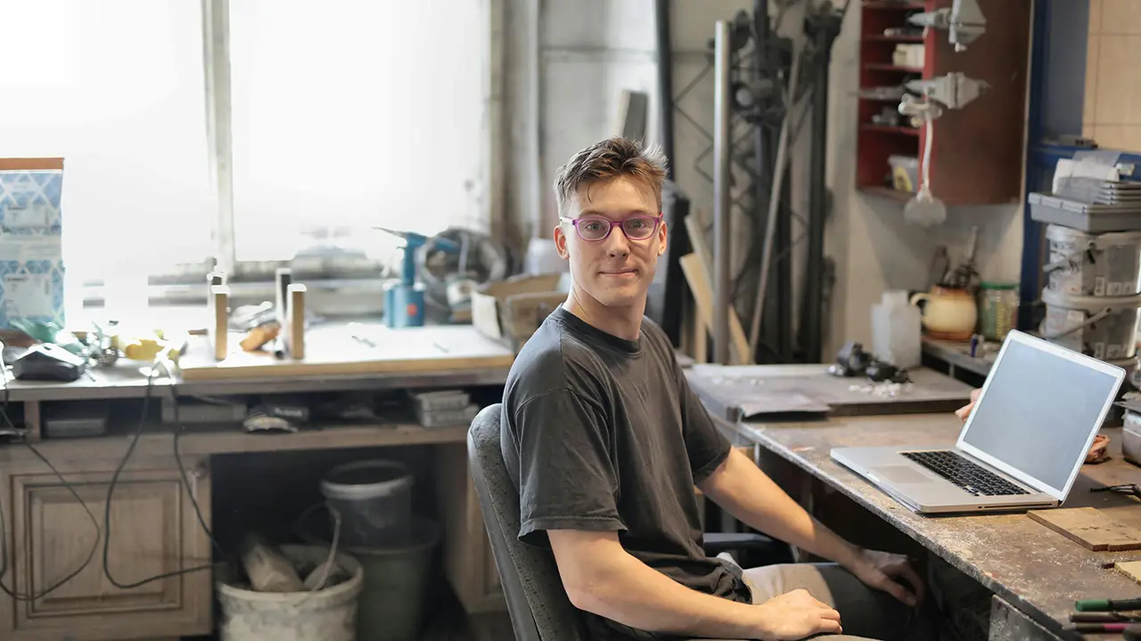Junger Handwerker mit Brille sitzt in einer Werkstatt an einem Schreibtisch und nutzt einen Laptop zur Arbeit.